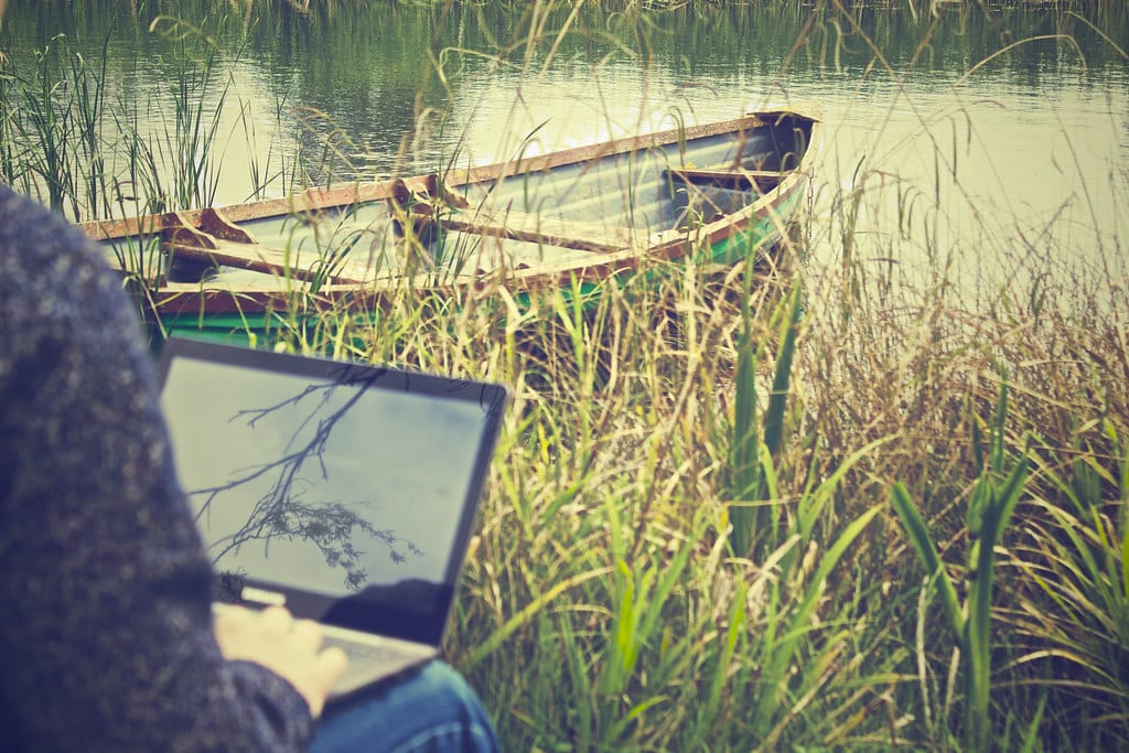 An image of a man working on his laptop outdoors beside a river and a small boat. Age inclusive brand communities can help oder people overcome their digital disadvantage and connect online