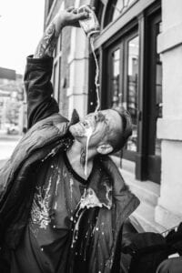 Black and white image of a man pouring beer from a can into his mouth from a height, beer spilling all over his face and clothes.