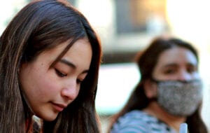 Image of two girls. One girl is in the foreground in focus, and the other is in the background wearing a face mask. 