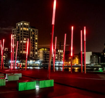 The red glowing angled light sticks of Grand Canal Square in Dublin Docklands at night, surrounded by green, polygon-shaped planters. High-rise buildings with lights turned on, can be seen in the background
