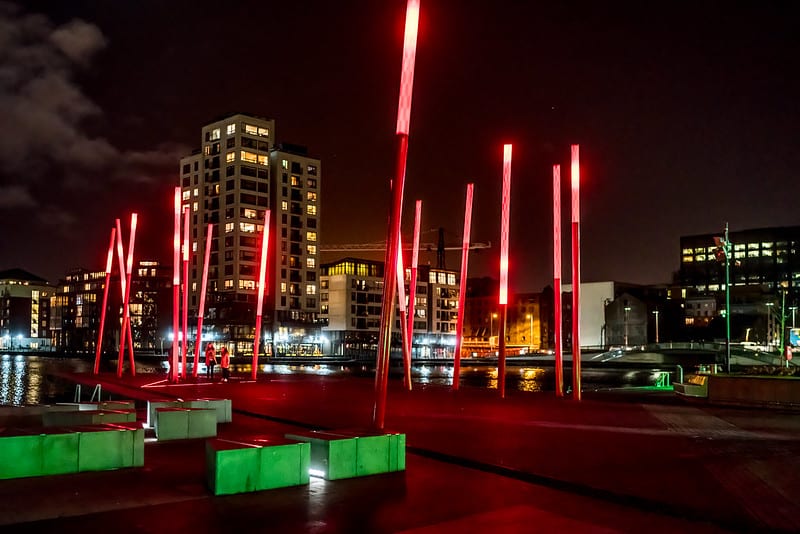 The red glowing angled light sticks of Grand Canal Square in Dublin Docklands at night, surrounded by green, polygon-shaped planters. High-rise buildings with lights turned on, can be seen in the background