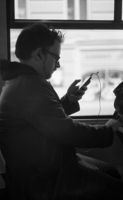This is a black and white image of a man looking at his phone screen while on a bus with earphones in. 