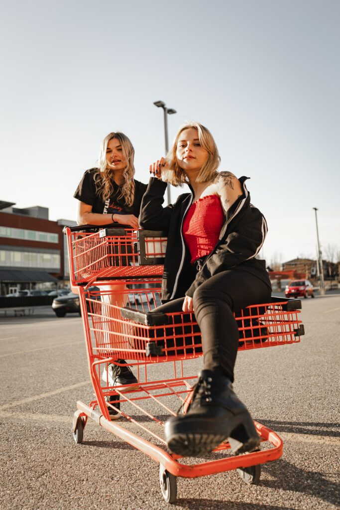 Two stylish teenage girls in the picture. One teenage girl in black jeans, red tube top, and black cardigan sitting in an orange shopping trolley while another teenage girl in a black tshirt stands behind the trolley 