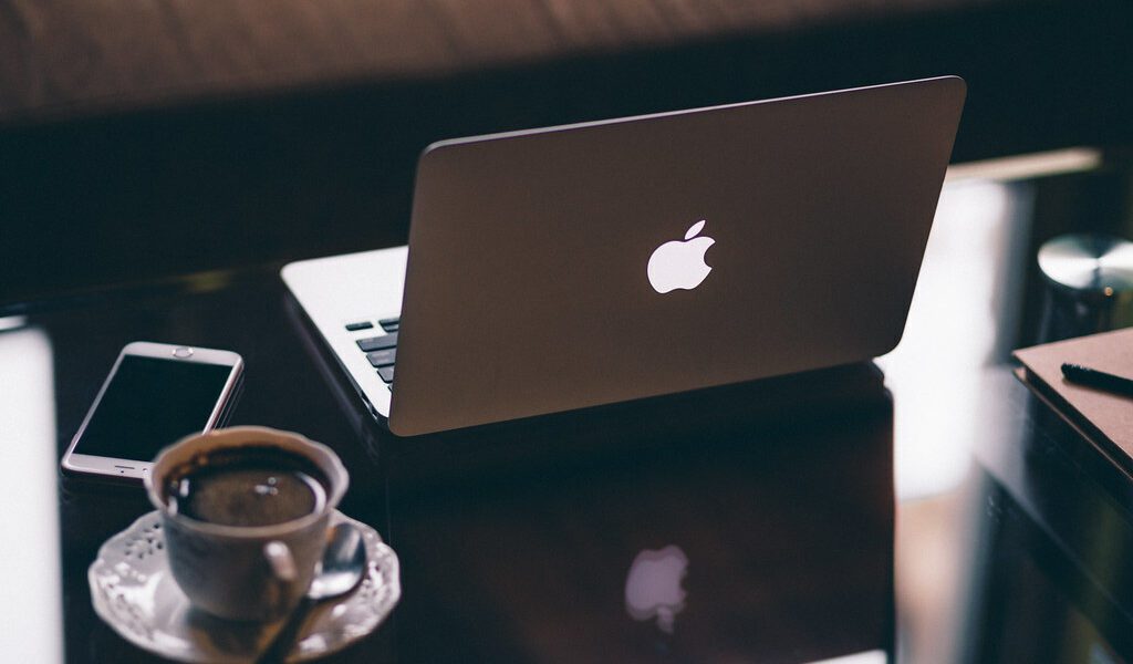 Image of a white apple mac book laptop in the middle of the image. On the left hand side of the image is a white coffee cup and saucer with black coffee inside and white smart mobile phone on a mirror effect desk reflecting each item.