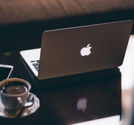 Image of a white apple mac book laptop in the middle of the image. On the left hand side of the image is a white coffee cup and saucer with black coffee inside and white smart mobile phone on a mirror effect desk reflecting each item.