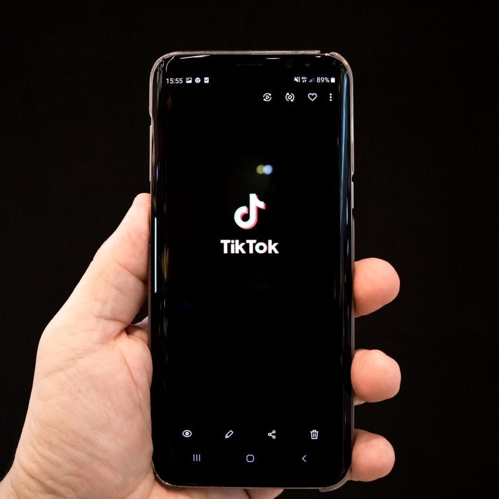 On a black background, a hand holds a phone displaying the digital app, TikTok