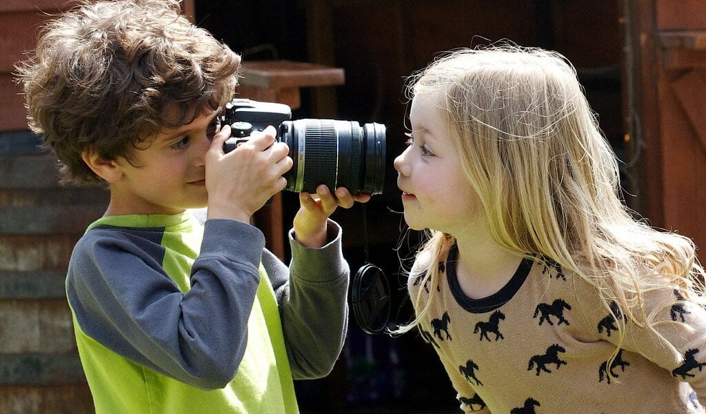 Image of young child using a camera to take a photo of another child, on a sunny day.
