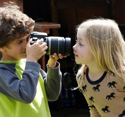 Image of young child using a camera to take a photo of another child, on a sunny day.