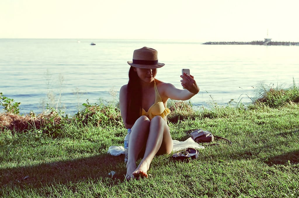 Girl sits in front of a lake taking a selfie. Social media selfie filters contribute to selfie dysmorphia