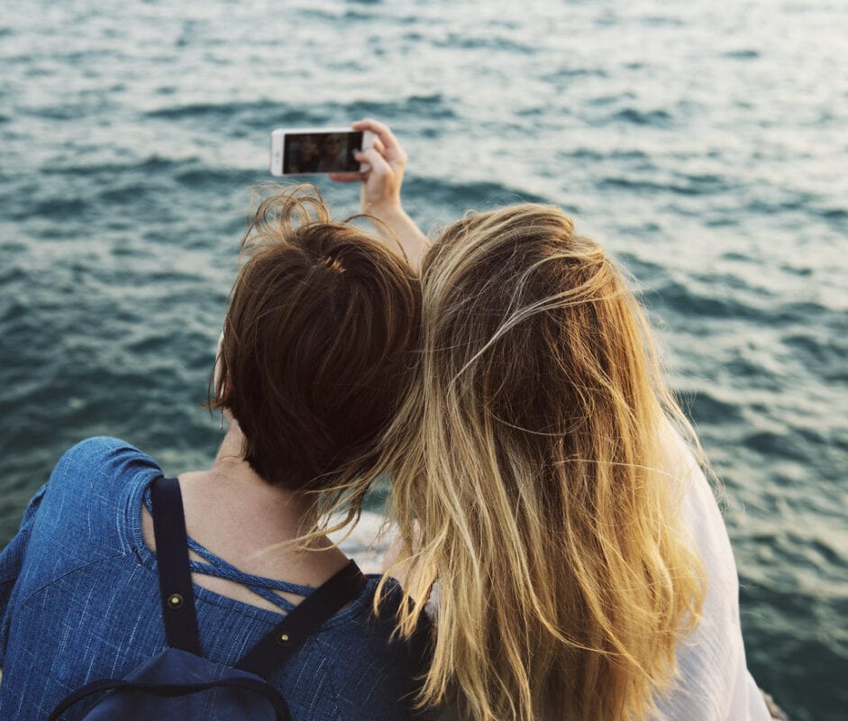 Two girls sit in front of the sea whilst taking a selfie