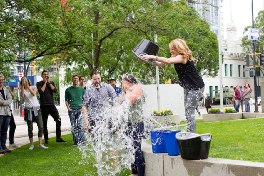 Group of smiling and laughing people watch as one woman dumps a bucket of ice water onto another woman's head.