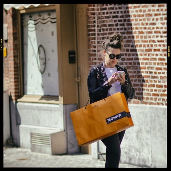 This image is of a person walking down the street with an orange shopping bag on their arm. They are holding their phone in both hands and looking down at it. They are wearing black jeans, a white top, a black leather jacket, sunglasses, and have their hair up in a bun.