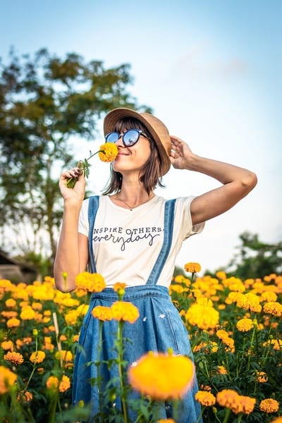 This is an image of an Instagram influencer promoting a chosen brand's clothes. Portraying that money can buy you happiness. She is wearing a blue denim dress: bottom half is denim along with straps; and top half is a white top shape with the words "inspire others everyday" printed on it. She is also wearing a hat and sunglasses, and is smelling a yellow flower with one hand, whilst the other is lifted to touch her hat. She is stood in a field of tall yellow flowers on a sunny day. There is a tree in the background, and a blue sky.