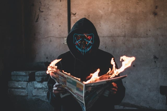 Person in mask and hoodie holding a burning fake news newspaper close-up photography